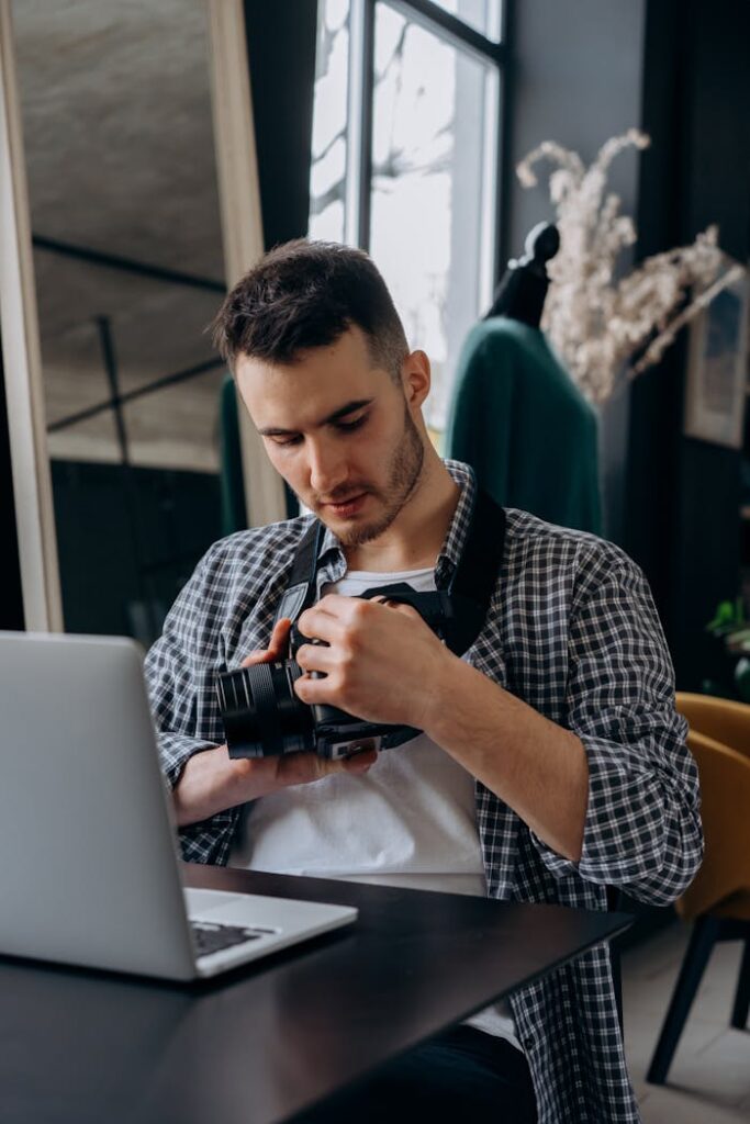 A young photographer in casual clothing examines a camera while working in a modern home office setting.
